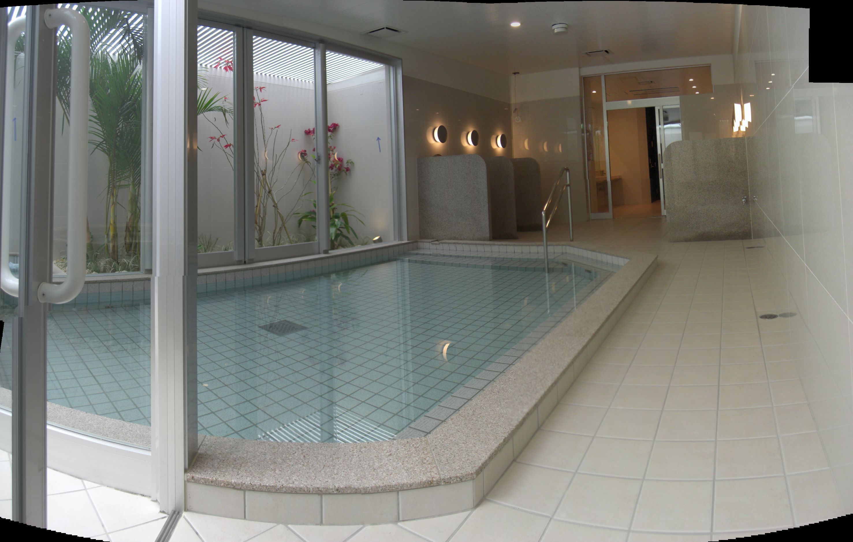 Una piscina con paredes de cristal transparente, rodeada de suelo y paredes con azulejos, con vistas a plantas de interior y una puerta que da a otra habitación.