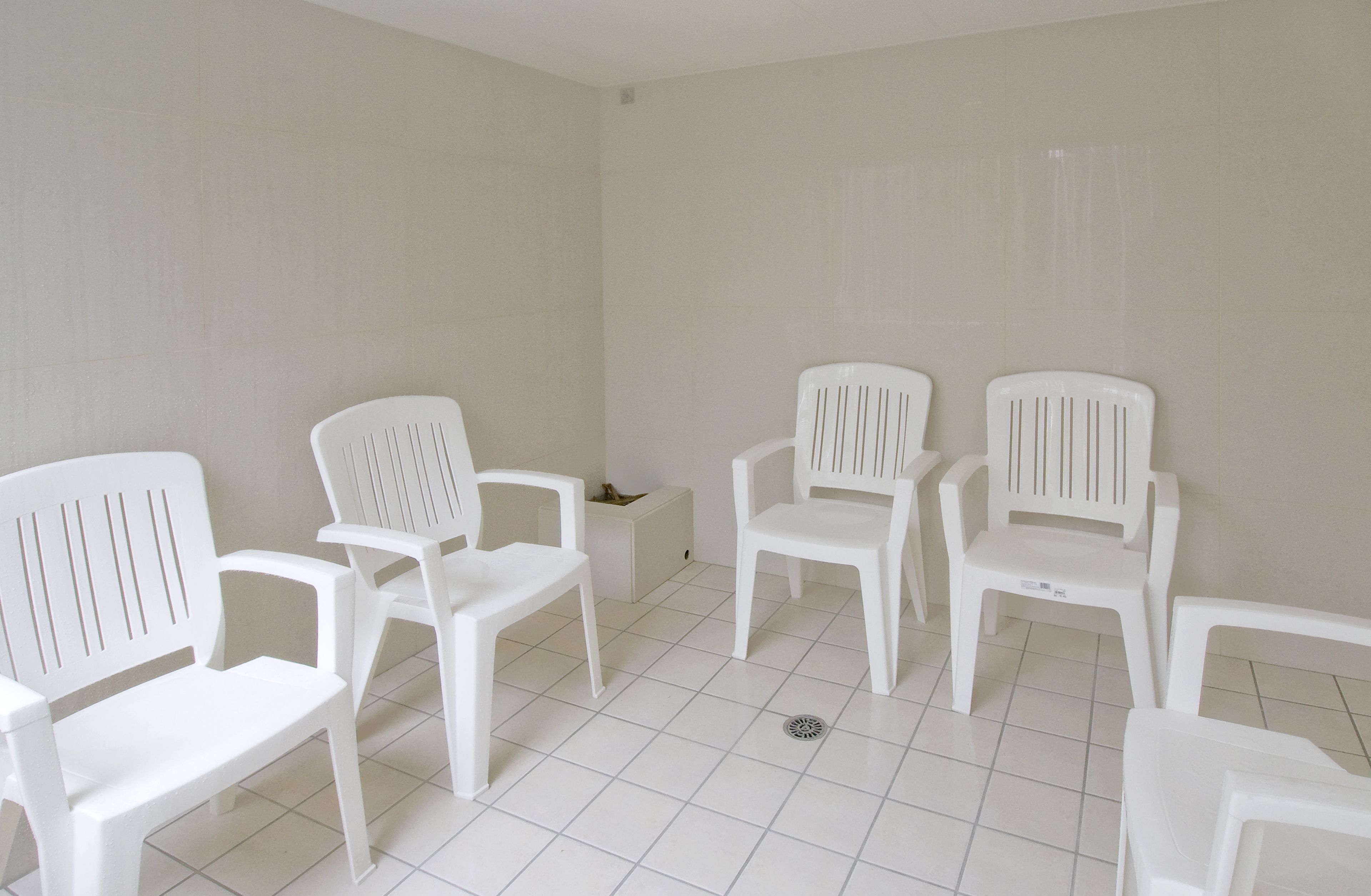 Una habitación con suelo y paredes de baldosas blancas, que contiene cinco sillas de plástico blanco dispuestas en semicírculo.