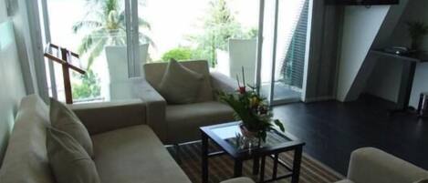 A modern living room with a sofa, a coffee table, and a TV. There is a large window offering a view of palm trees and a building.