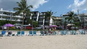 Una playa con sillas de descanso y sombrillas, palmeras y edificios de varios pisos al fondo.