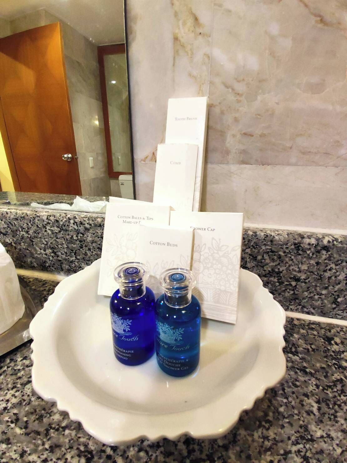 Un tocador de baño con encimera de mármol, un lavabo blanco y artículos de aseo personal como gorro de ducha, bastoncillos de algodón y peine.