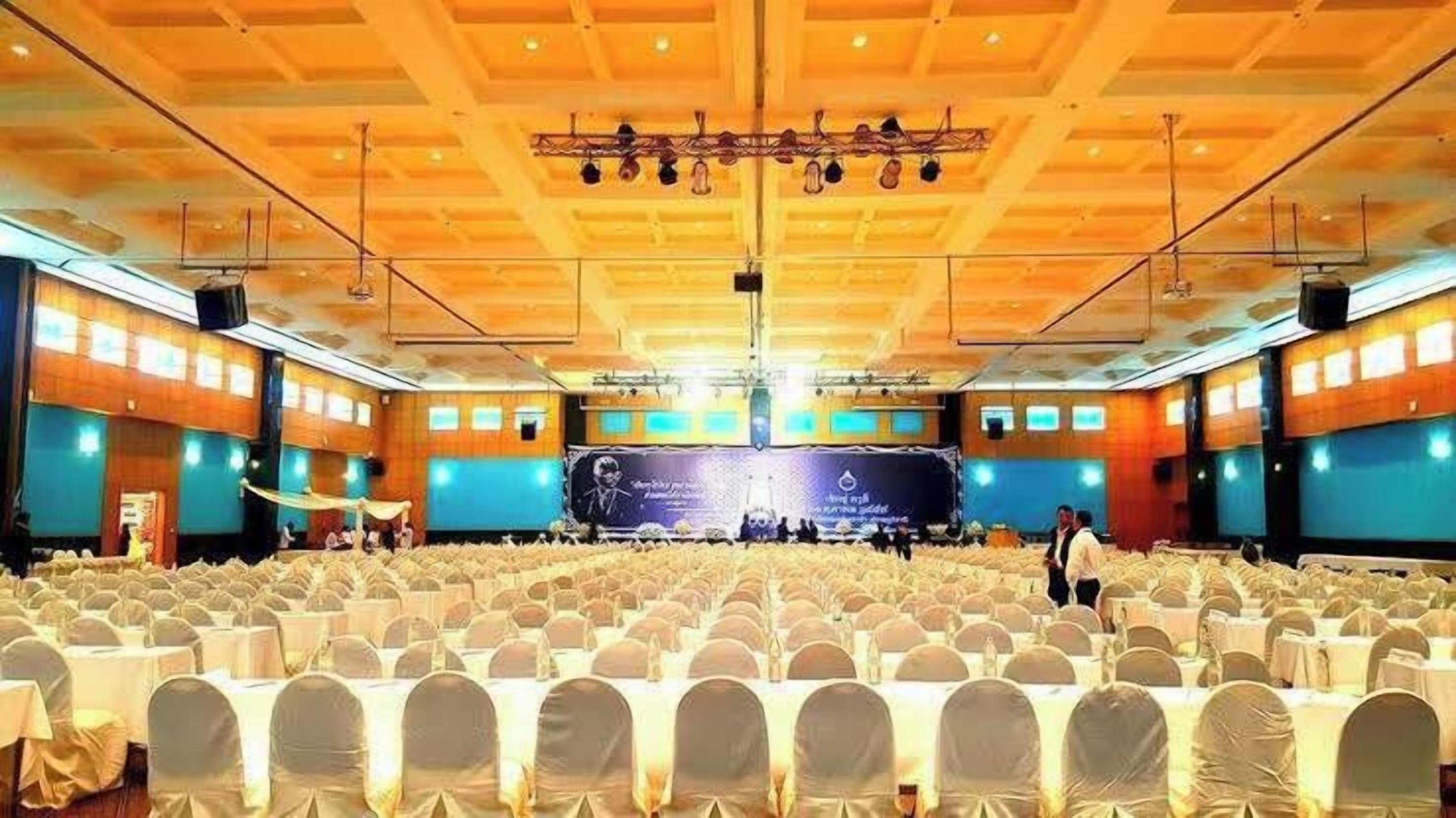 A large hall with rows of tables and chairs, a stage with a banner, and a high ceiling with lighting fixtures.
