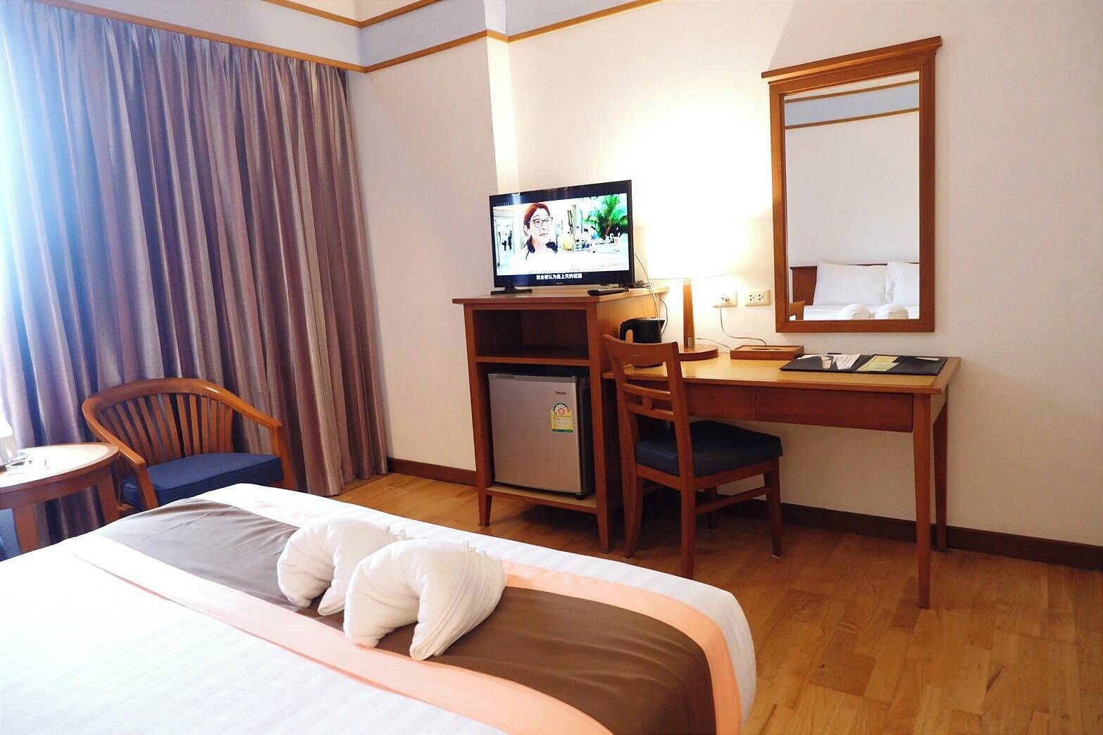 A hotel room with a bed, a desk with a computer, a chair, a small refrigerator, and a large mirror.