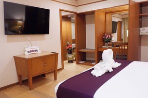 Una habitación de hotel con una cama, un televisor montado en la pared, un tocador de madera, un espejo y un baño visible a través de una puerta abierta.