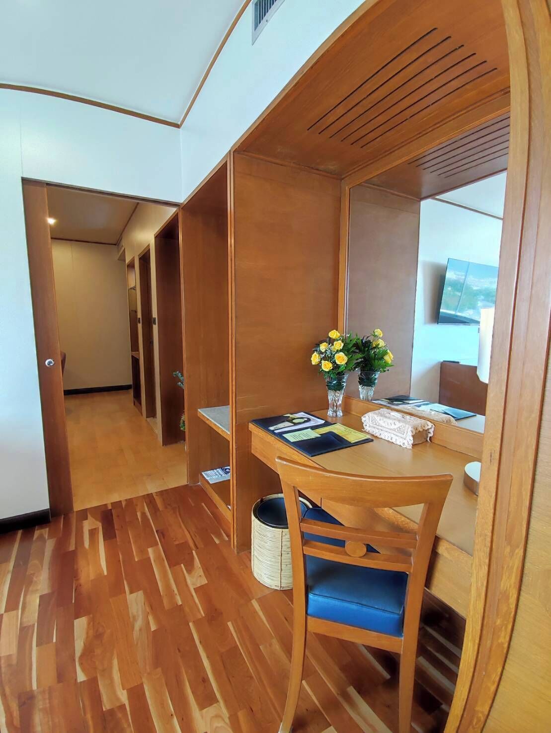 A hotel room with a wooden desk, a chair, a small table, and a vase with flowers.