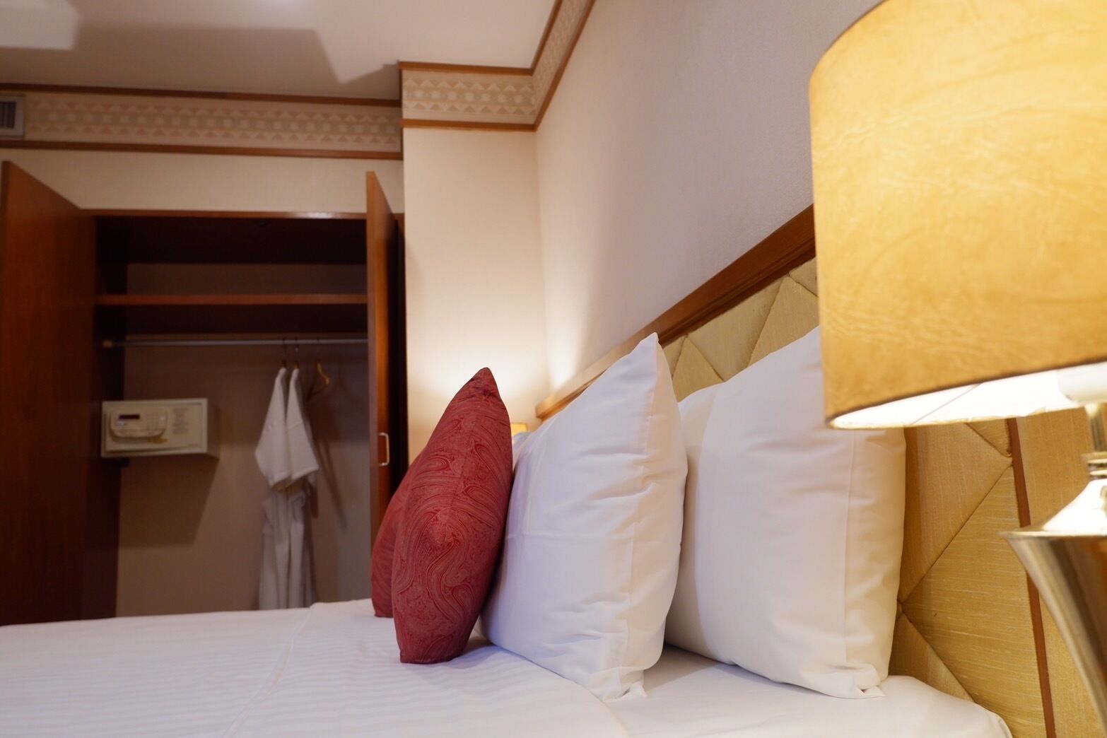 Une chambre d’hôtel avec un lit, un oreiller rouge et blanc, une lampe et une armoire contenant une robe et un panneau de commande.