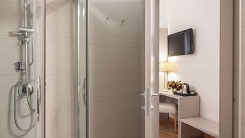 A modern bathroom with a glass shower enclosure, a mounted television, and a bedside table with a lamp and decorative items.