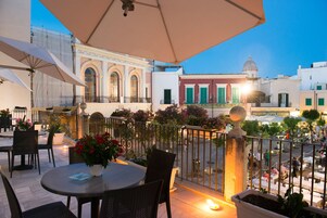 Una terrazza all'aperto con tavoli e sedie rotonde, una pianta in vaso e vista sulla città al crepuscolo.