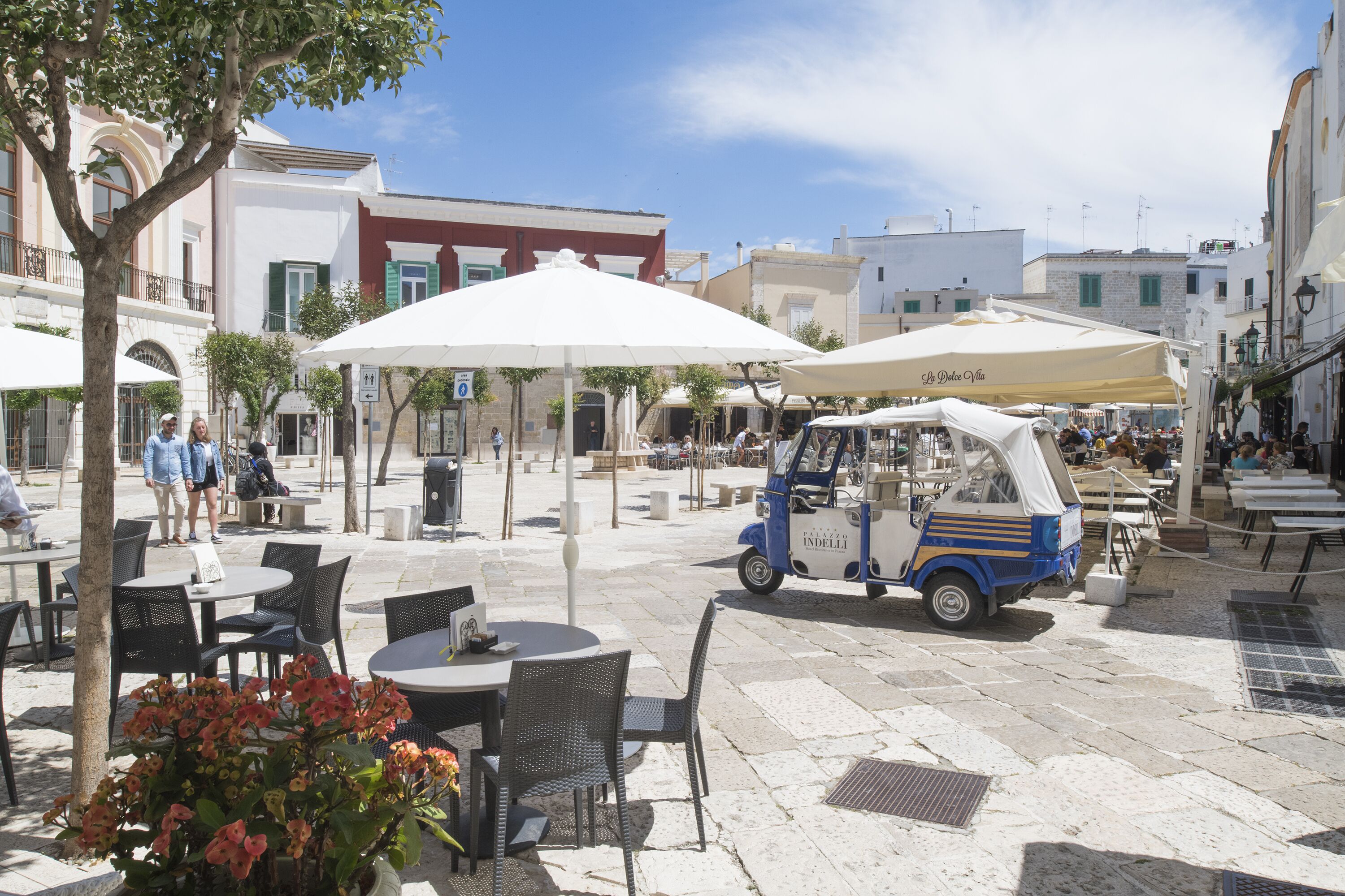 Un área de café al aire libre con mesas y sillas, un vehículo pequeño azul y blanco, y edificios al fondo.