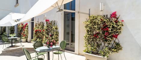 Un patio al aire libre con mesas y sillas, un paraguas blanco y plantas en macetas con flores.