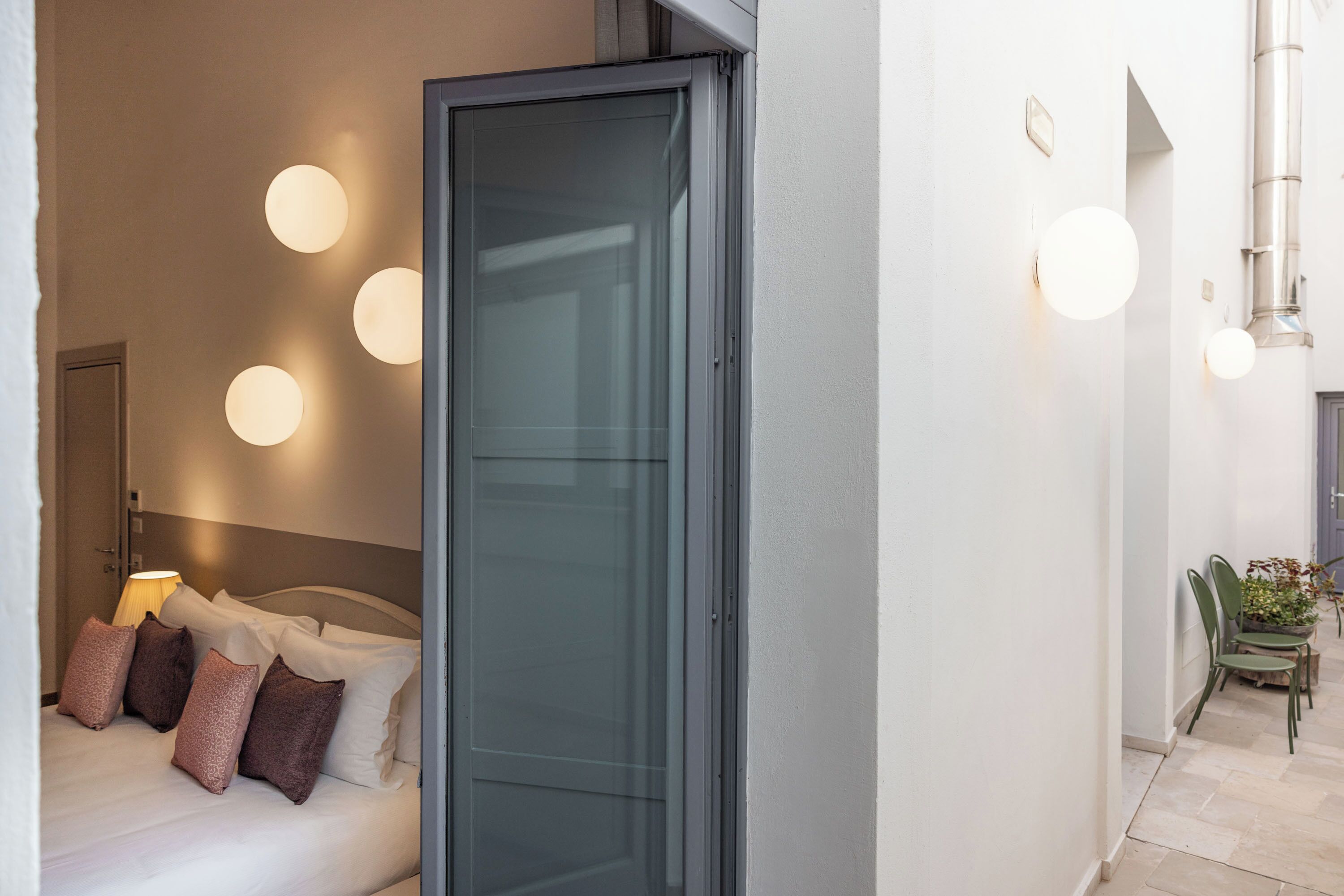 A hotel room with a bed, pillows, a nightstand, and a lamp. There is a sliding door leading to a hallway with chairs and a plant.