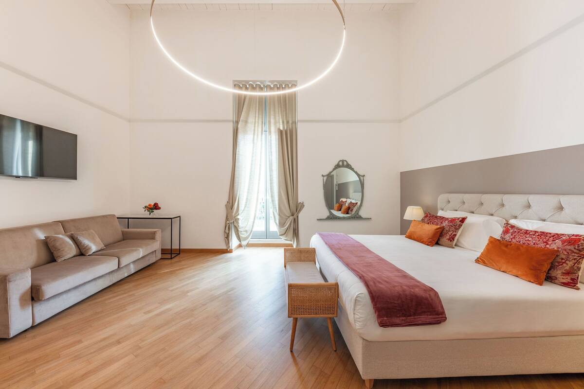 Una habitación de hotel moderna con una cama grande, un sofá, una pequeña mesa con un florero y un espejo.