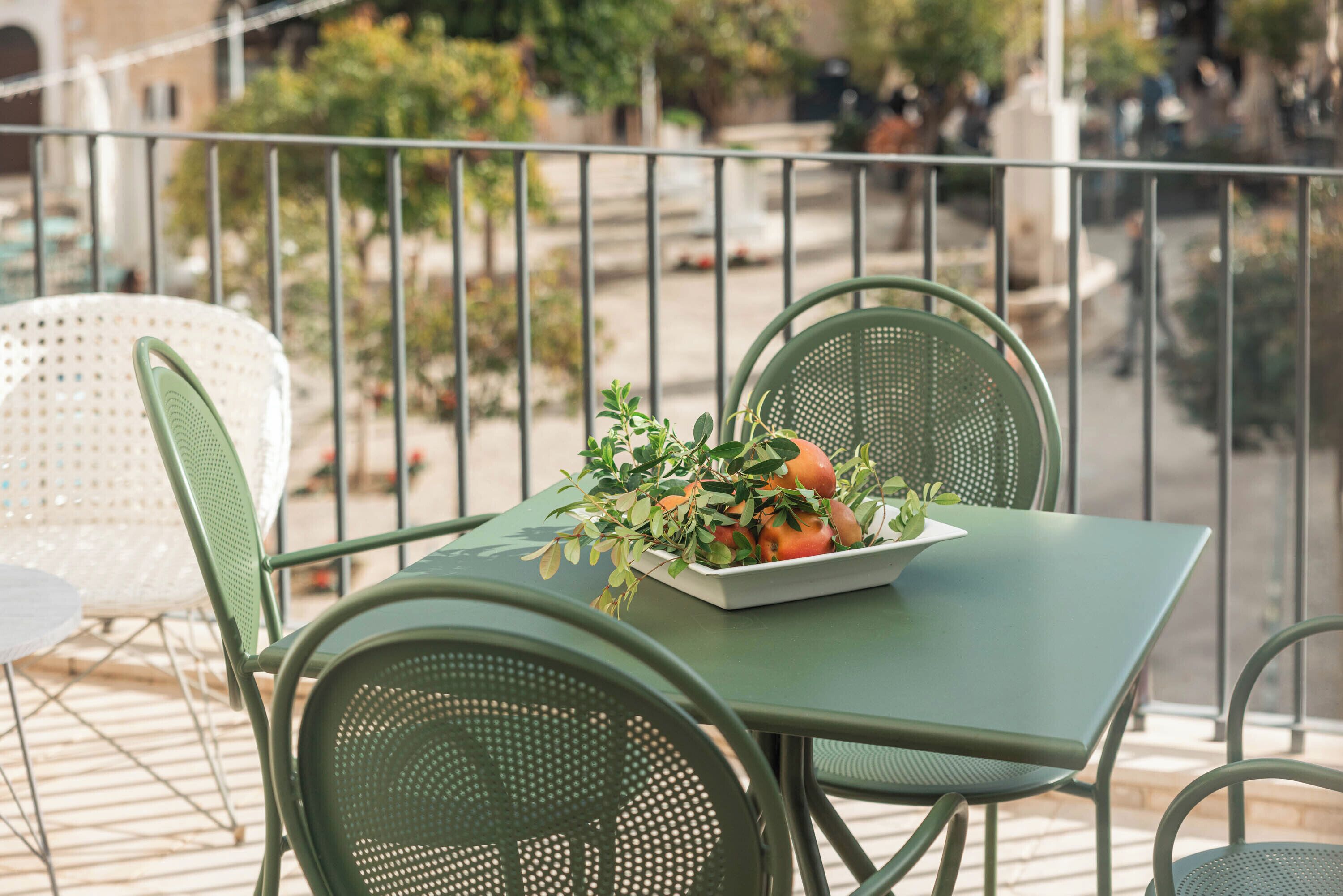 A balcony with a green metal table and chairs, a white table, and a bowl of fruit and greenery.