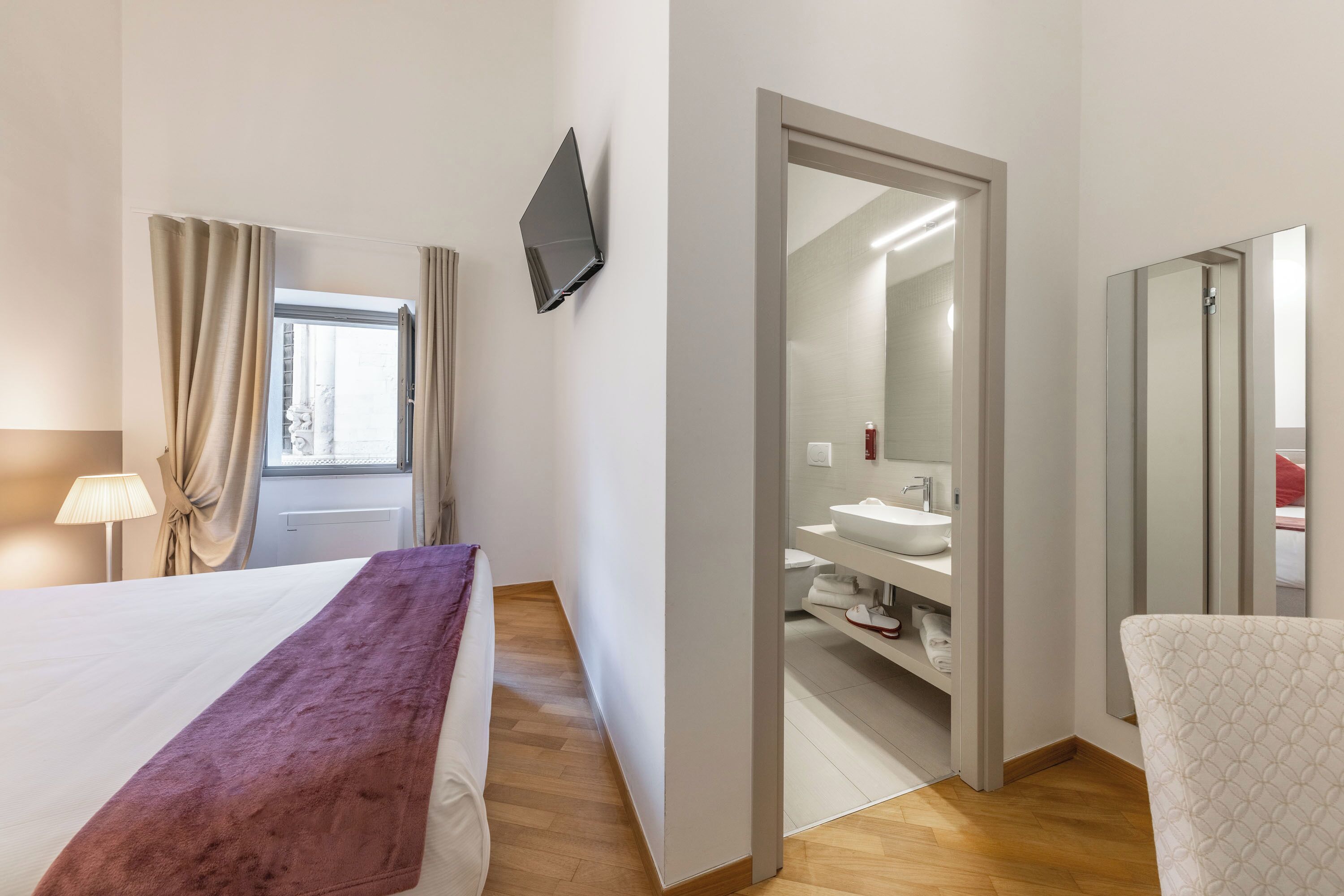 A hotel room with a bed, a TV mounted on the wall, a bathroom with a sink and mirror, and a window with curtains.