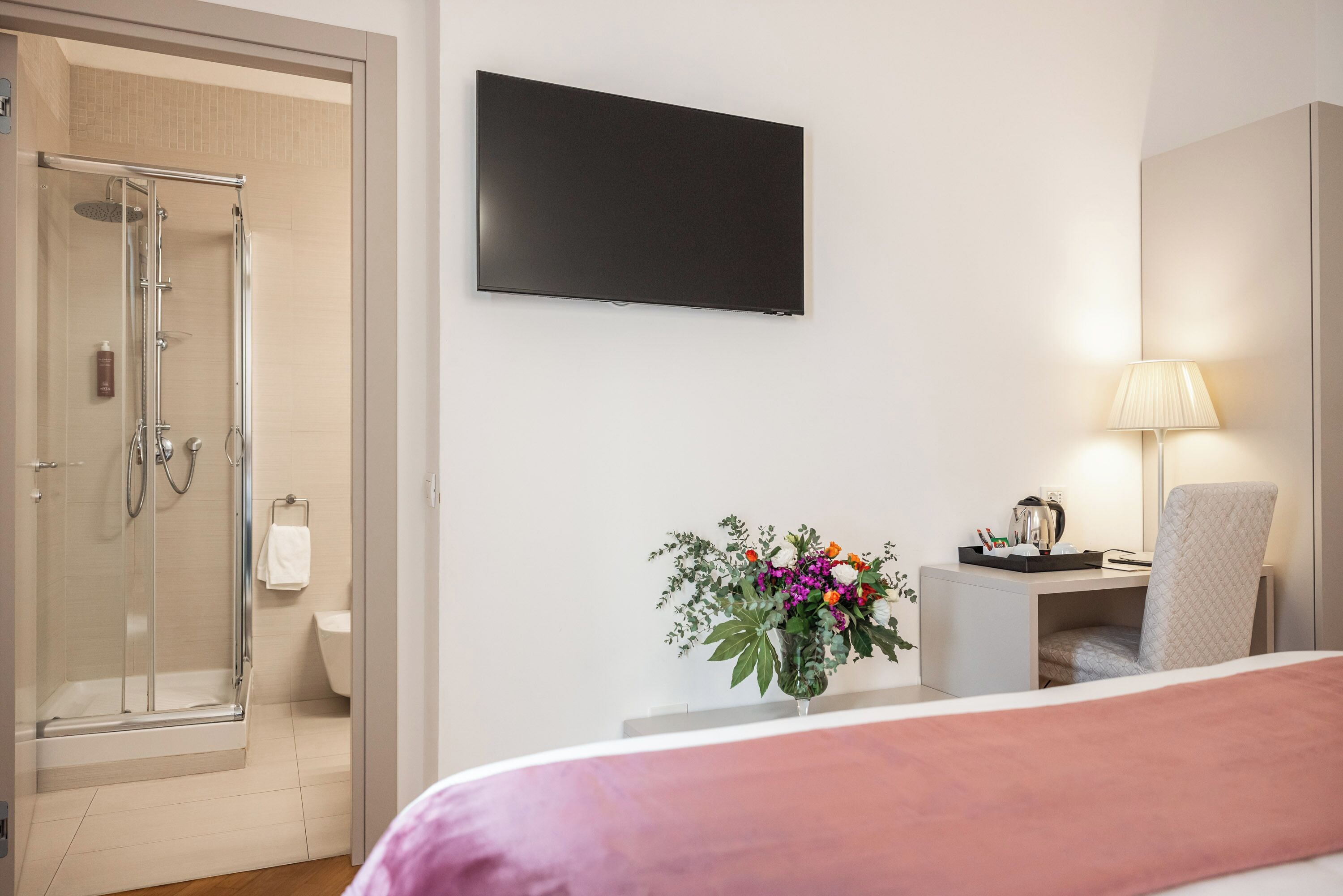 Een hotelkamer met een bed, een bureau met lamp, een televisie, een douche en een bloevas.