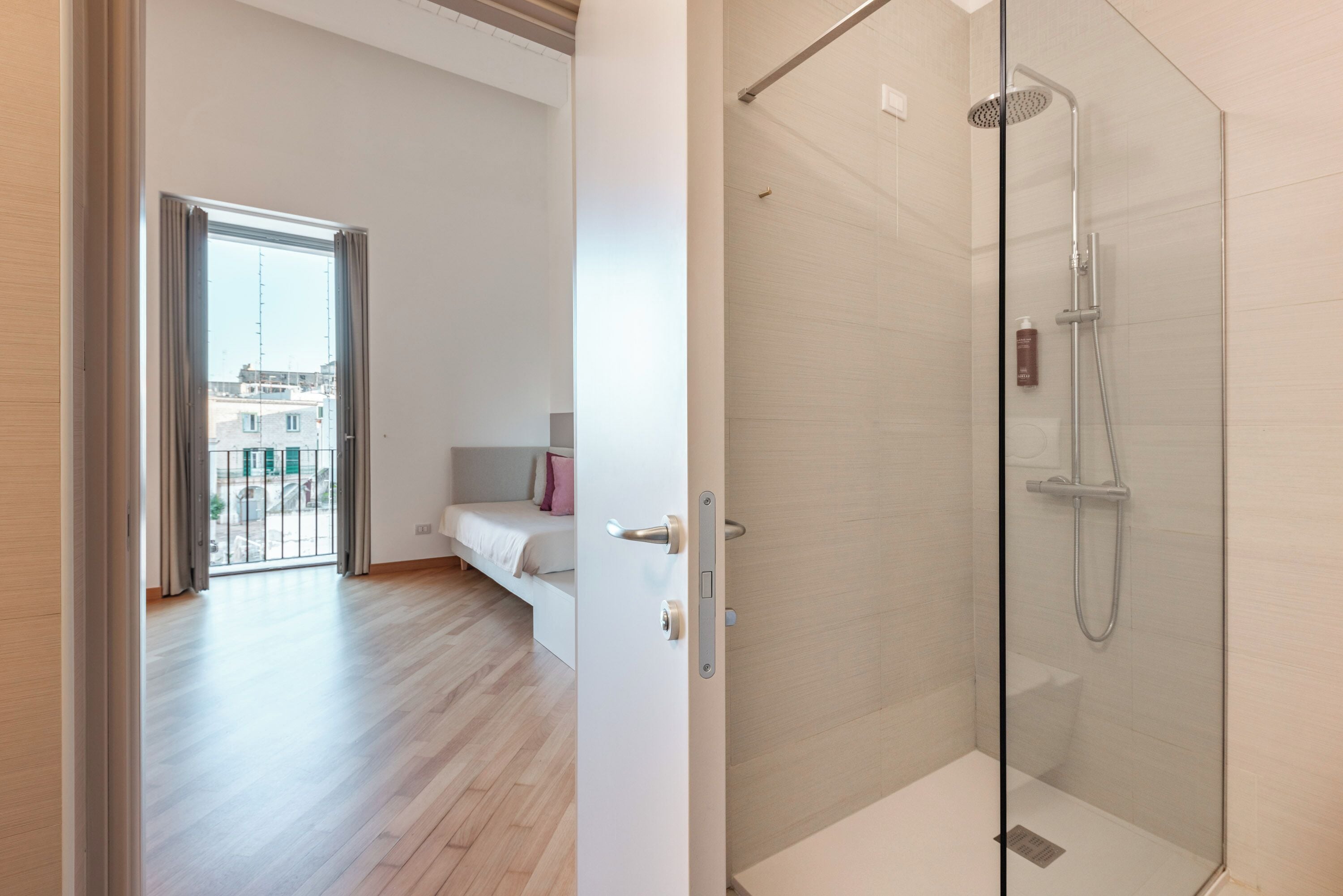 A modern hotel room with a glass-enclosed shower, a balcony with a bed, and a view of the cityscape.