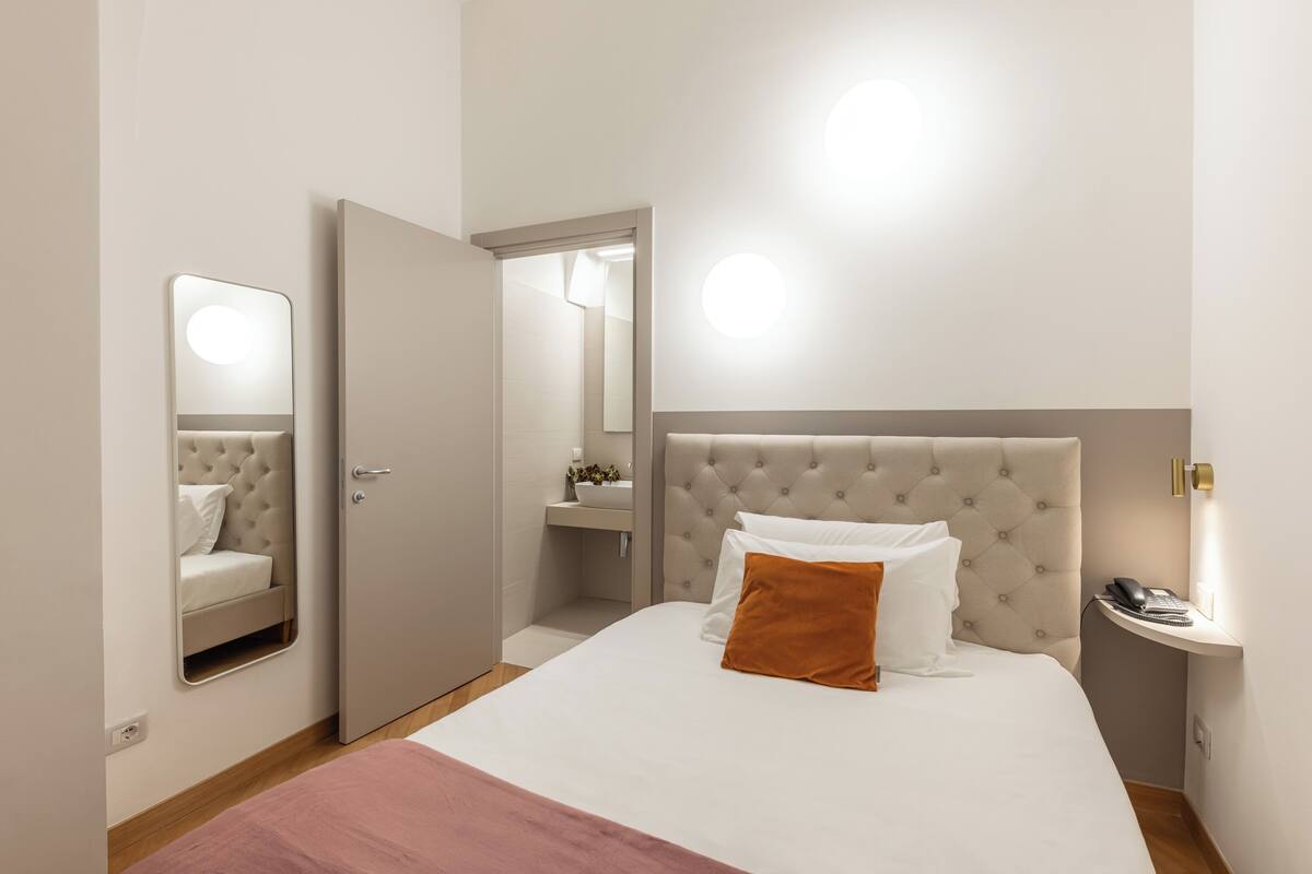 Un dormitorio con una cama grande, una mesita de noche, un espejo y un baño visible a través de una puerta abierta.