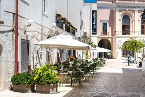 An outdoor café with tables and chairs, potted plants, and a cobblestone street. - Palazzo Indelli (Monopoli)