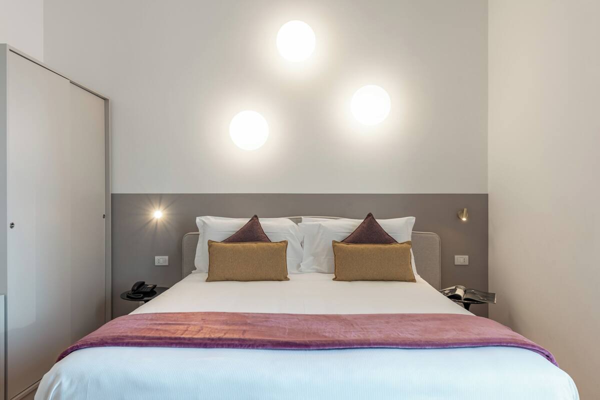 Habitación de hotel con una cama, dos almohadas, una mesita de noche con un teléfono, y una lámpara fijada a la pared.