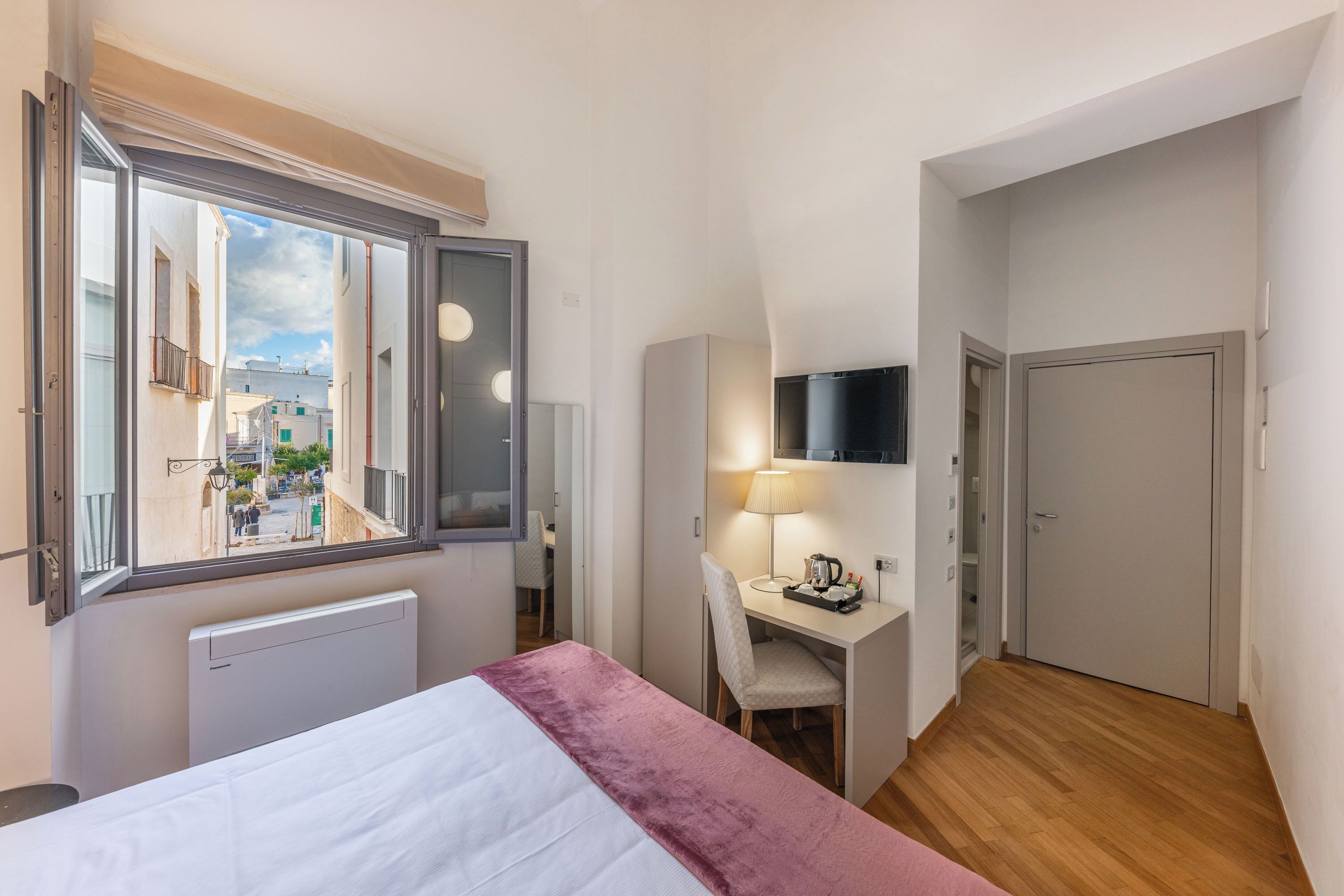 A hotel room with a bed, a desk with a chair, a TV mounted on the wall, and a view of the street through a window.