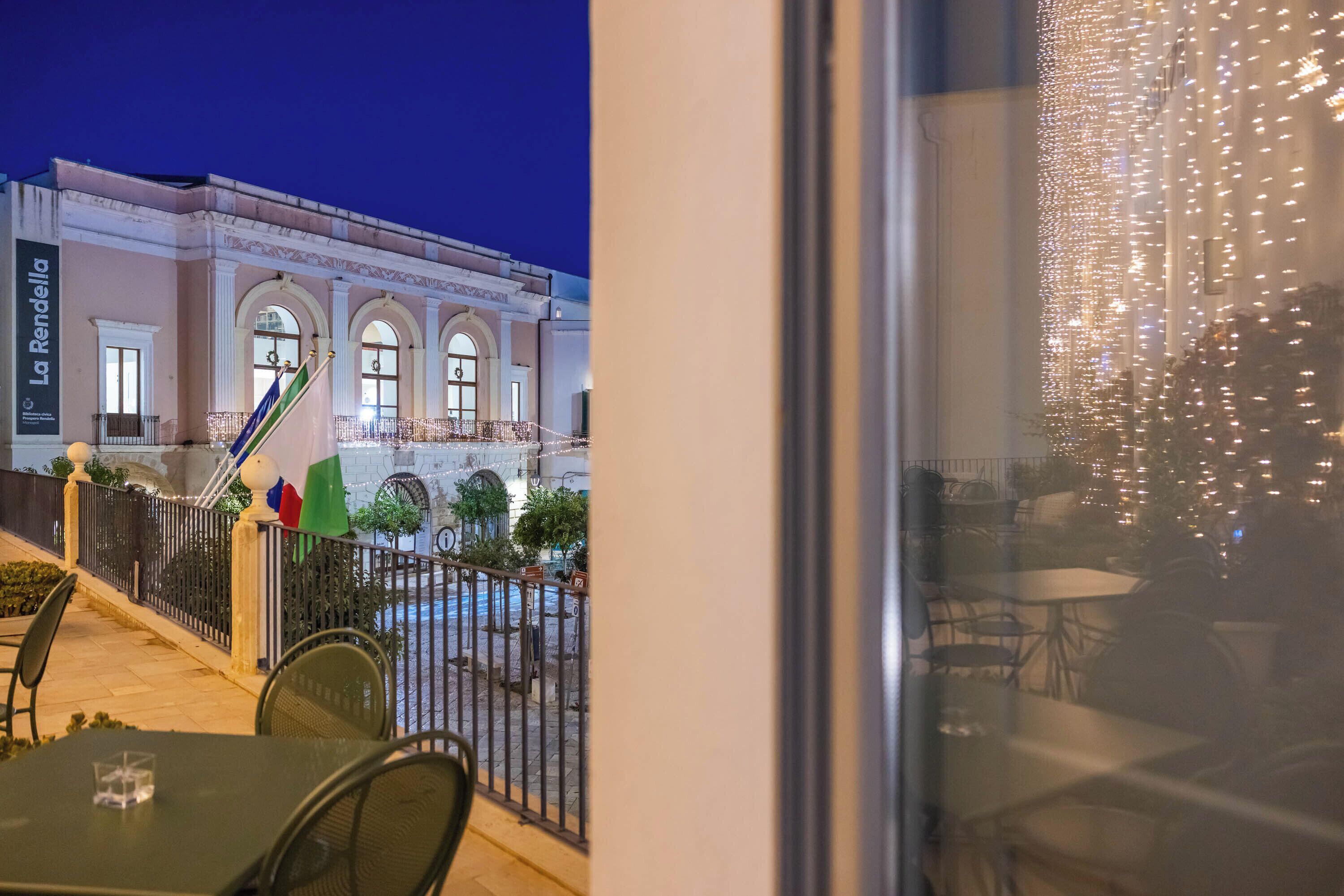 Un'area esterna con tavoli e sedie, una bandiera, e un edificio con finestre ad arco e un cartello con su scritto 'La Renda'.