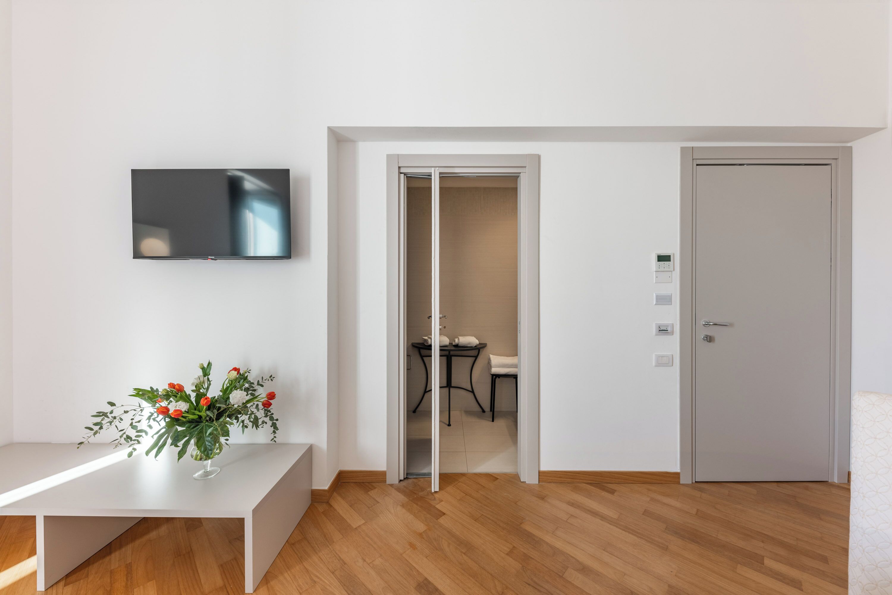 A modern room with a flat-screen TV mounted on the wall, a white bench with a floral arrangement, and a door leading to a bathroom.