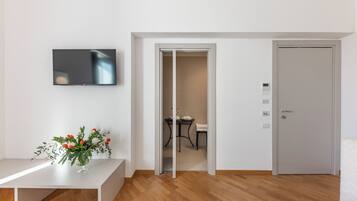 Quarto moderno com TV de tela plana na parede, um banco branco com arranjo floral e porta que dá para um banheiro.