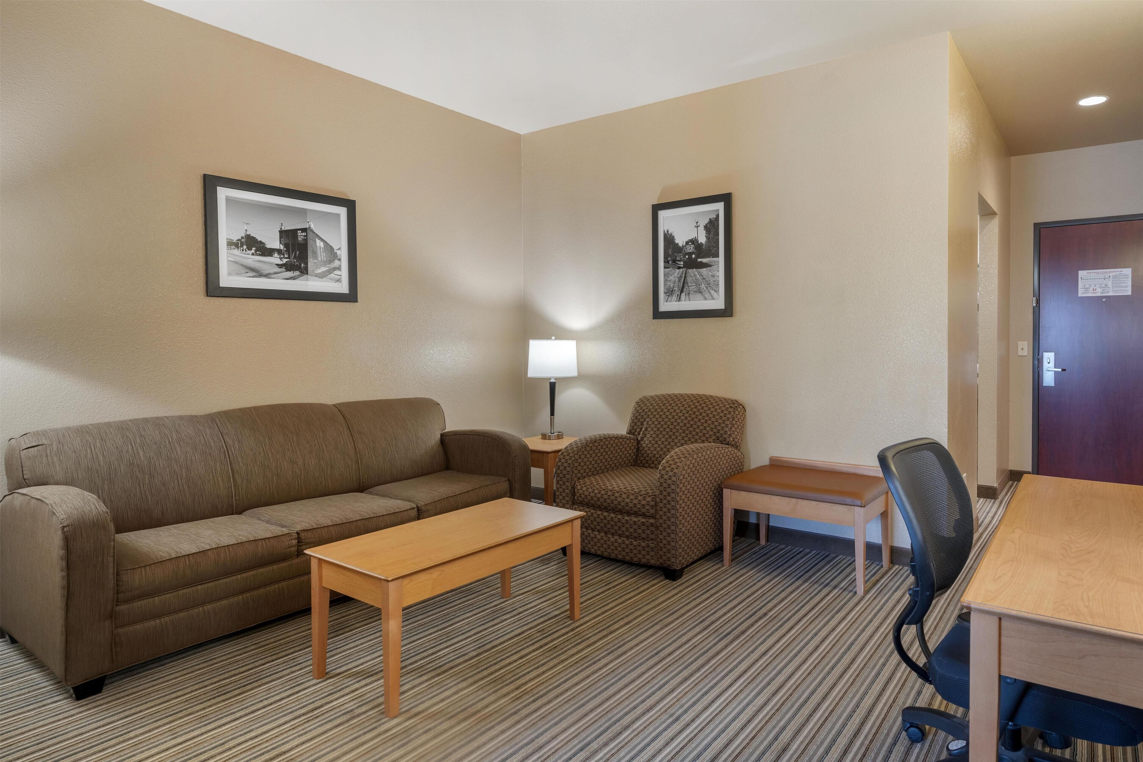 A hotel room with a sofa, armchair, small table, lamp, and a desk with a chair.