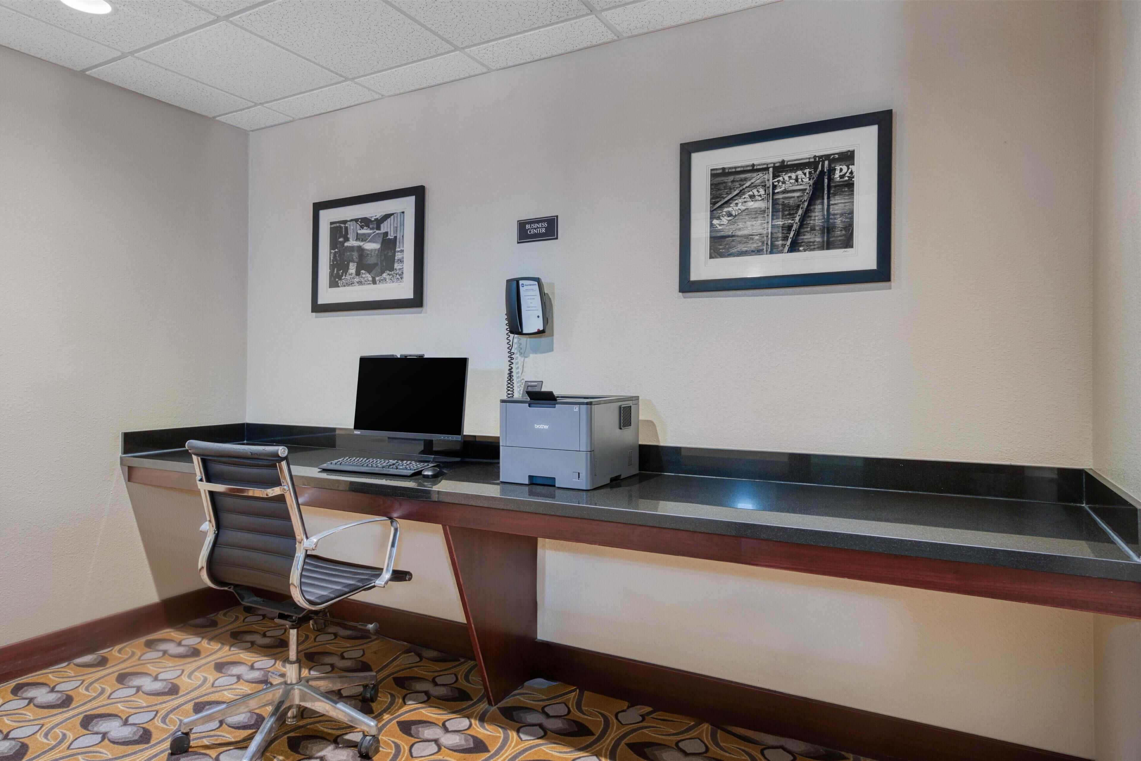 A hotel room with a desk, chair, computer, printer, and two framed pictures on the wall.