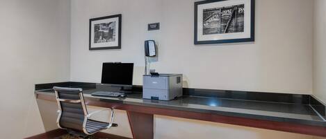 A hotel room with a desk, chair, computer, printer, and two framed pictures on the wall.