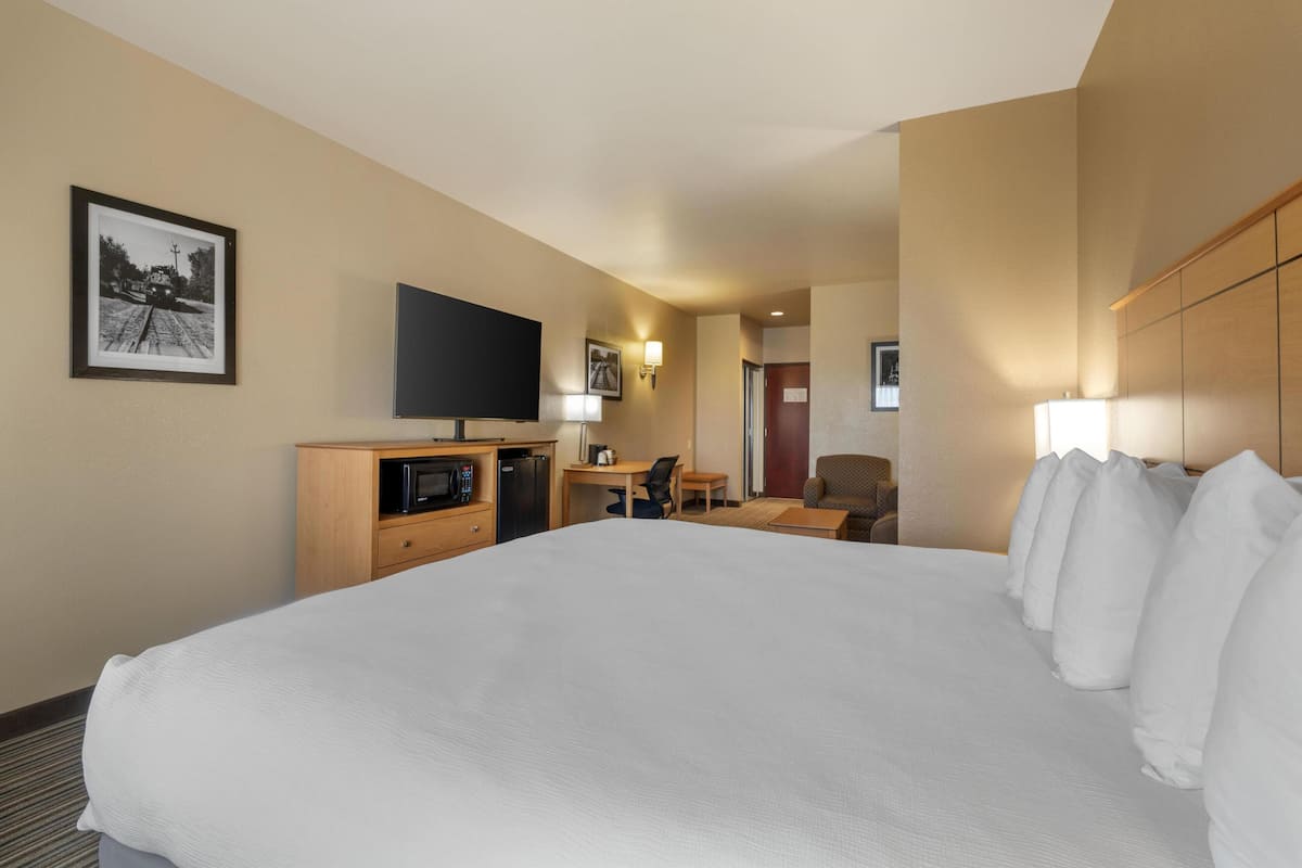 Ein Hotelzimmer mit einem großen Bett, einem Schreibtisch mit Stuhl, einem Flachbildfernseher, einer Mikrowelle und einem kleinen Tisch mit Lampe.