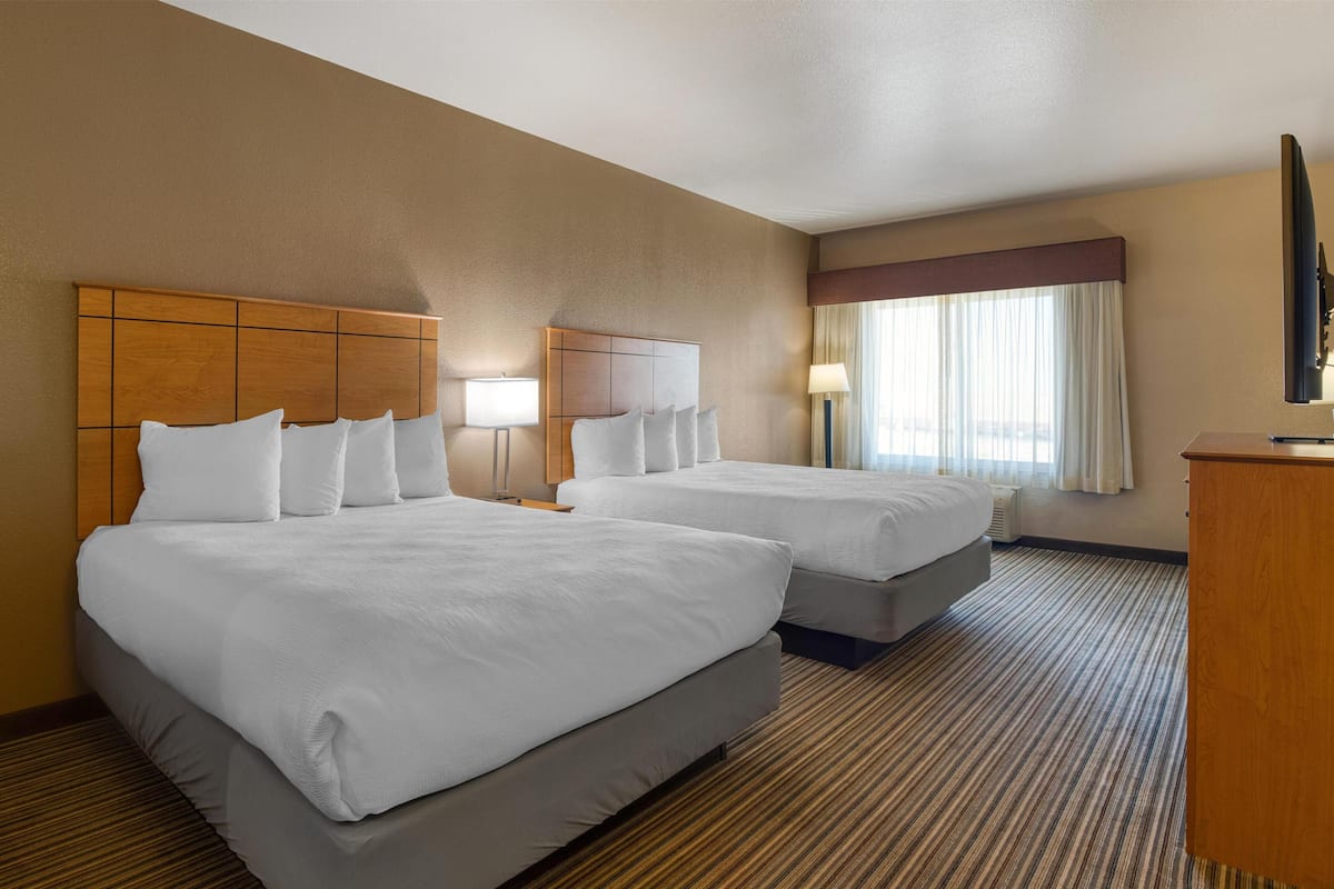 Ein Hotelzimmer mit zwei Betten, einem Holzkopfteil, einem Flachbildfernseher und einem großen Fenster mit Vorhängen.