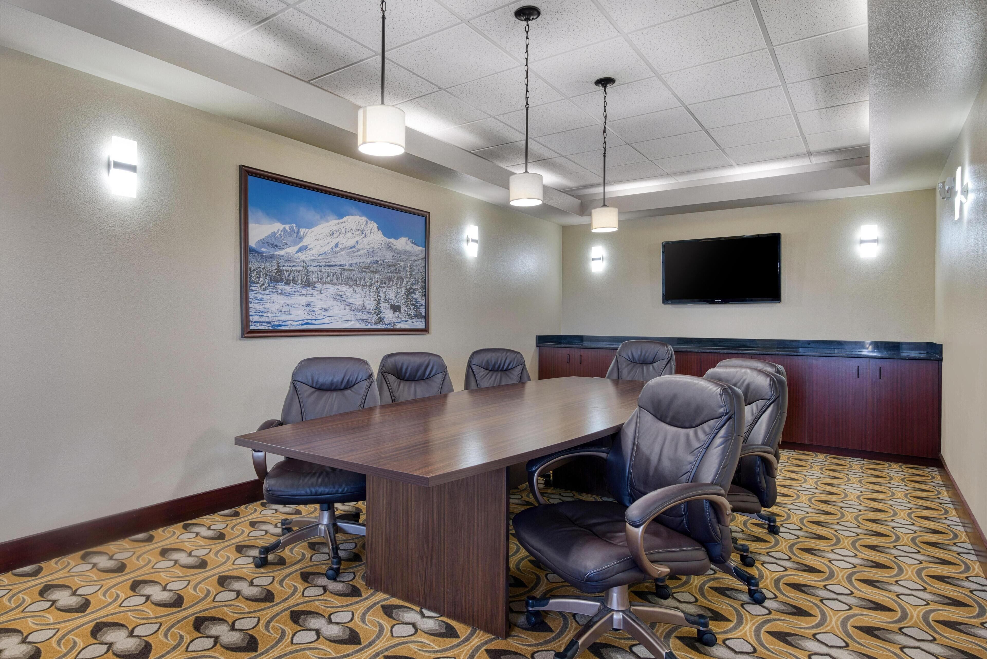 A conference room with a large table, six chairs, a mounted TV, and a painting of a snowy mountain landscape.