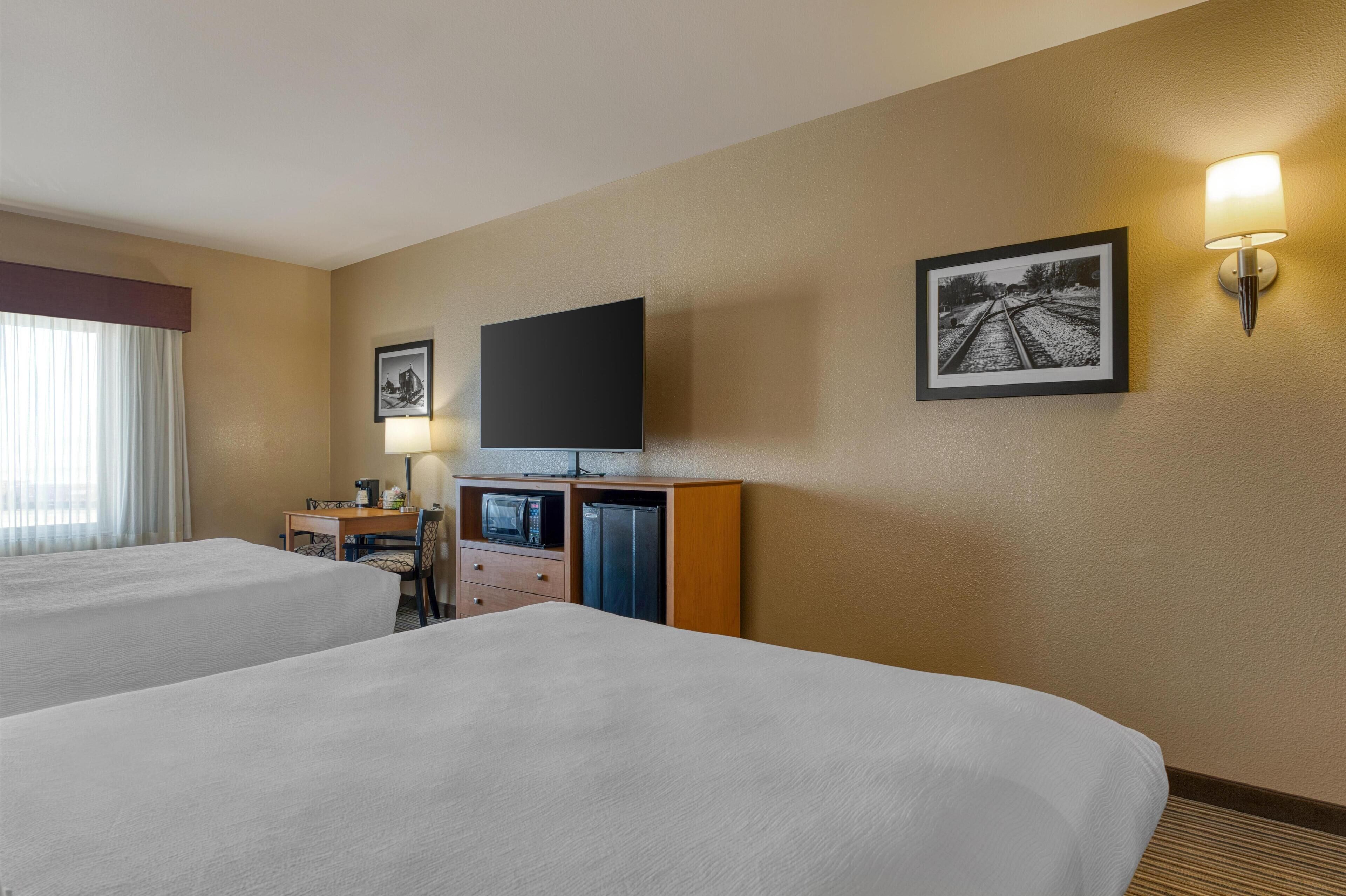 A hotel room with two beds, a desk with a chair, a microwave, a refrigerator, a flat-screen TV, and a wall-mounted lamp.