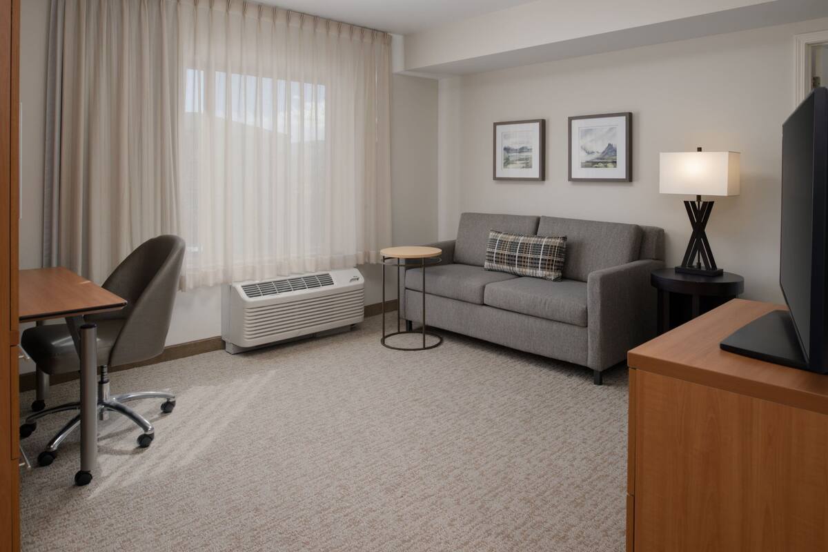 Een hotelkamer met een grijze bank, een kleine ronde tafel, een staande lamp, een televisie, een bureau met een stoel, en een raam met dunne gordijnen.