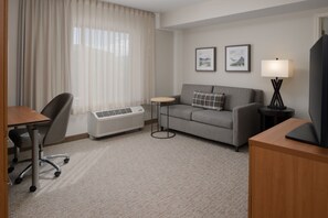 Une chambre d’hôtel avec un canapé gris, une petite table ronde, un lampadaire, une télévision, un bureau avec une chaise et une fenêtre avec des rideaux légers.
