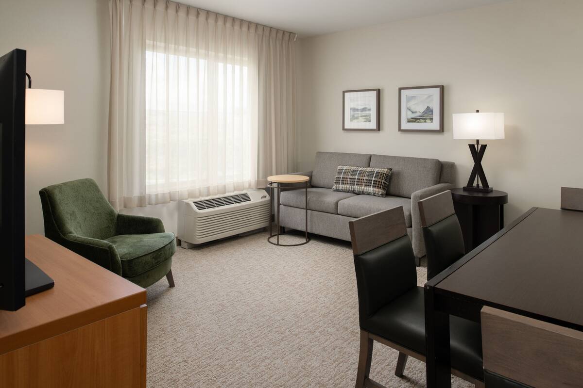 Een hotelkamer met een groene fauteuil, een grijze bank, een klein rond tafeltje, een televisie, een lamp en twee ingelijste foto’s aan de muur.
