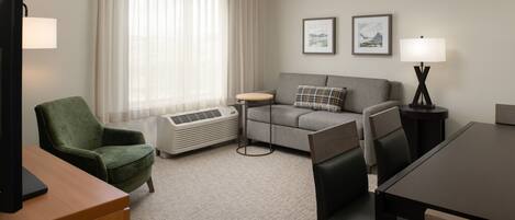 Habitación de hotel con un sillón verde, un sofá gris, una pequeña mesa redonda, un televisor, una lámpara y dos cuadros en la pared.