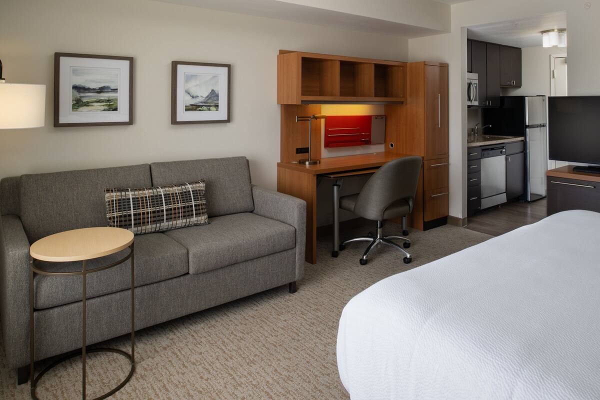 Een hotelkamer met een grijze bank, een kleine ronde tafel, een bureau met stoel, een bed, een televisie en een kitchenette.