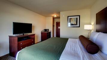 Habitación de hotel con cama, televisor sobre un soporte de madera, escritorio con lámpara y un cuadro en la pared.