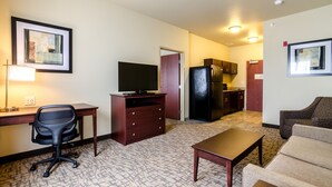 Ett kompakt hotellrum med ett litet kök, ett skrivbord med en stol, en soffa och en tv.