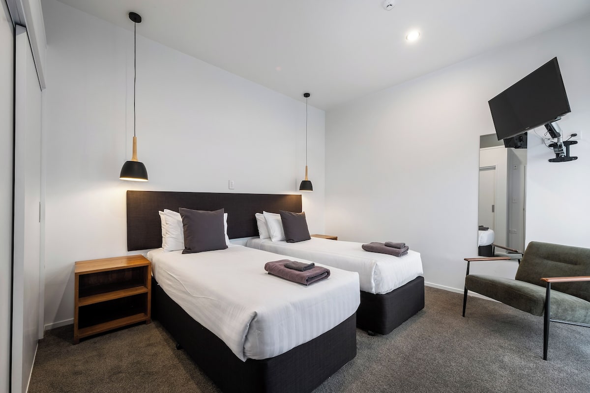 Une chambre d’hôtel moderne avec deux lits, une télévision, une chaise et une petite table de chevet en bois.