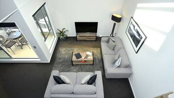A modern living room with a grey sofa, a coffee table, and a TV. There is a dining area with a table and chairs visible through an open door.