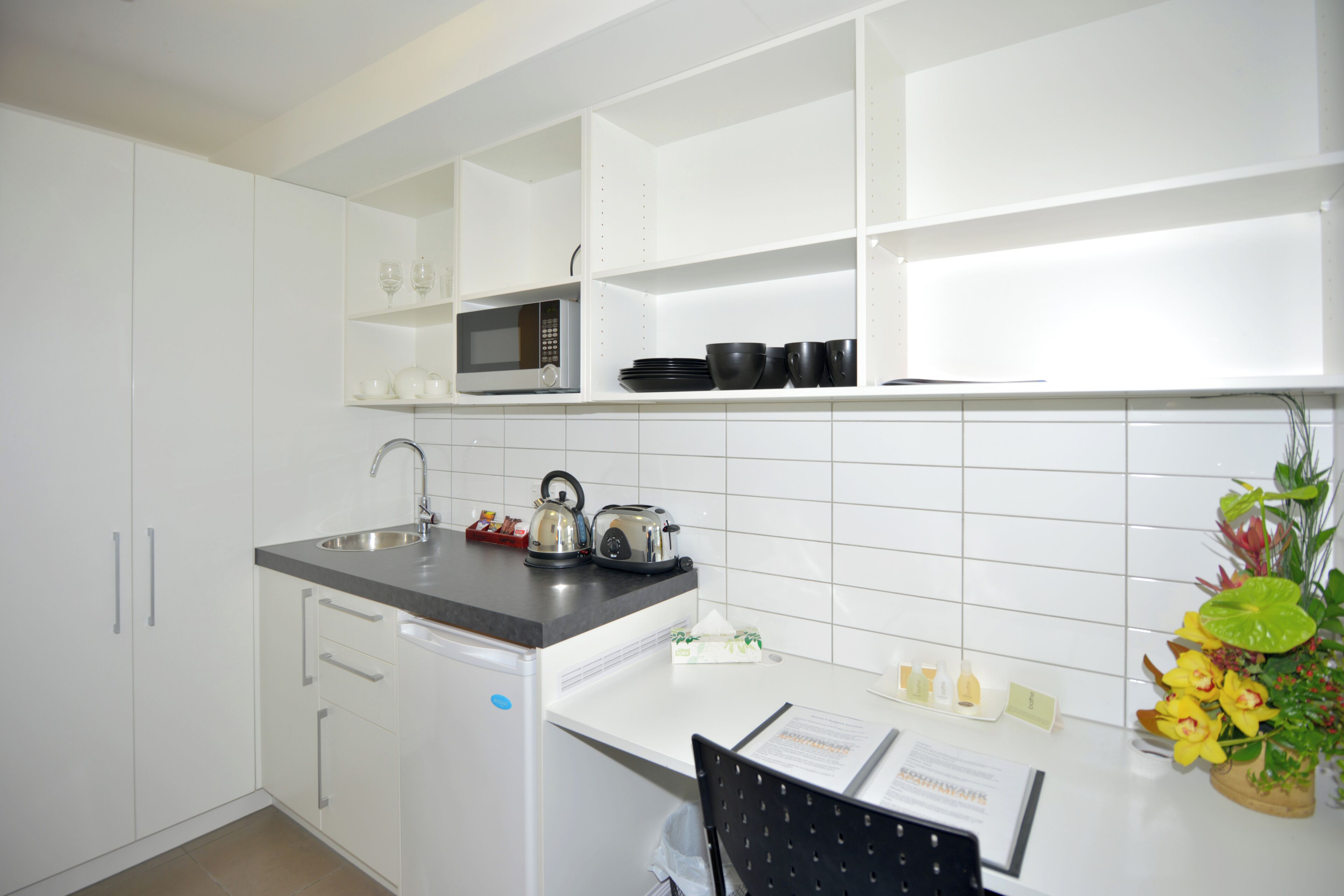Una cocina moderna con armarios blancos, un lavamanos de acero inoxidable, un microondas y un hervidor en la encimera. Hay una silla con un menú y una planta pequeña a la derecha.