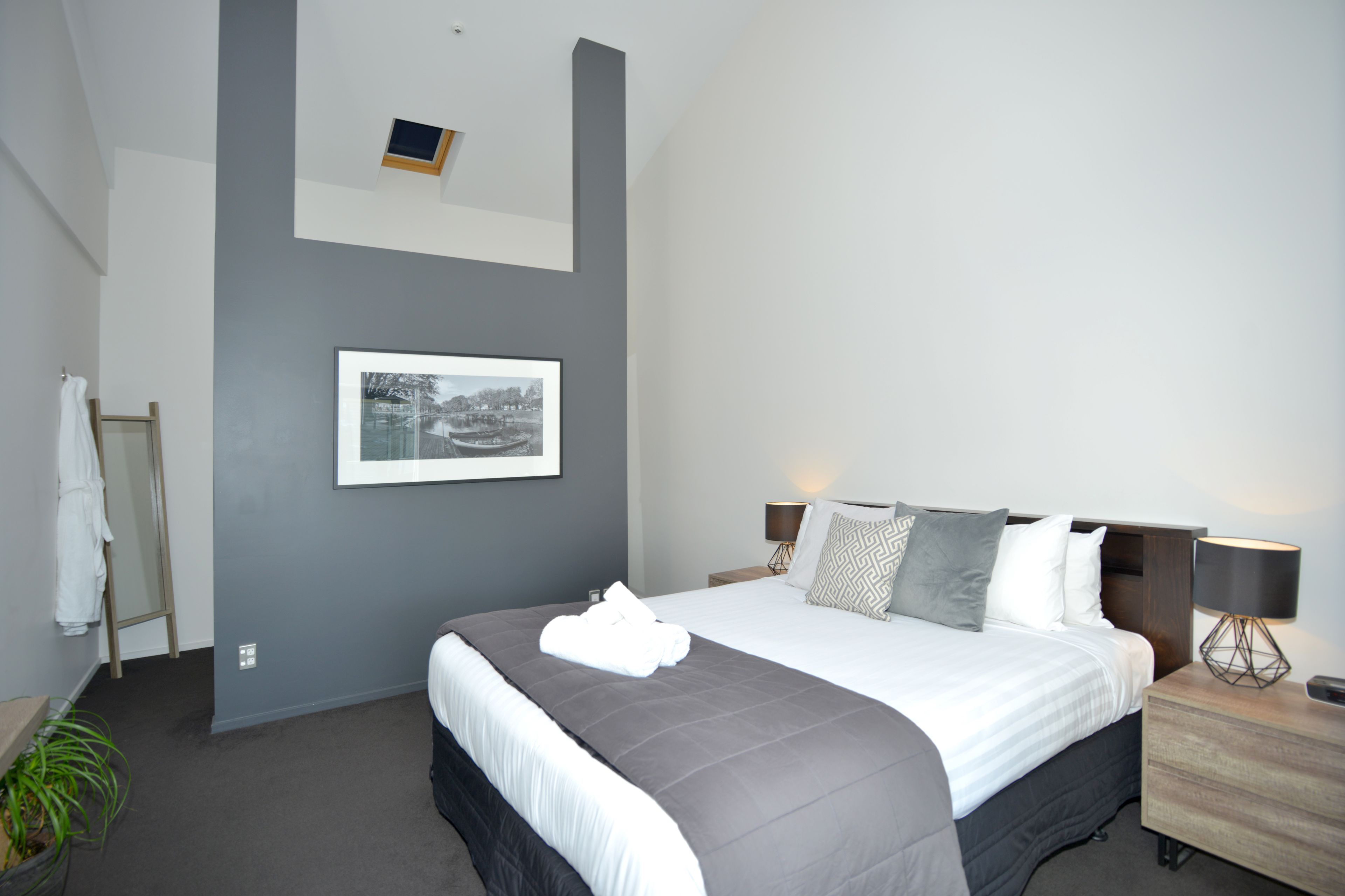 A modern hotel room with a large bed, a bedside table with a lamp, a framed picture on the wall, and a small plant.
