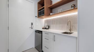 A compact kitchen with white cabinetry, a built-in dishwasher, a stainless steel sink, and wooden shelves above the countertop.