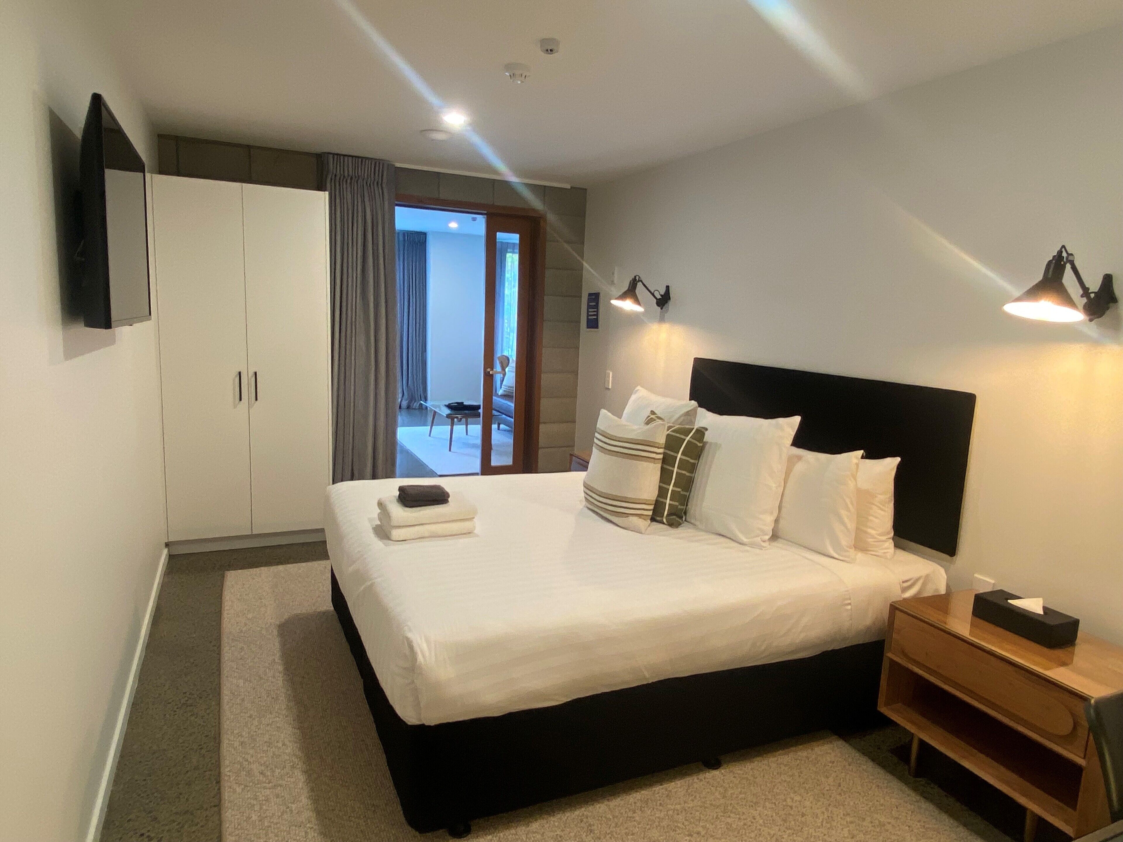 Ein modernes Hotelzimmer mit einem großen Bett, einem hölzernen Nachttisch, einem an der Wand befestigten Fernseher und Blick auf den Balkon durch die Tür.