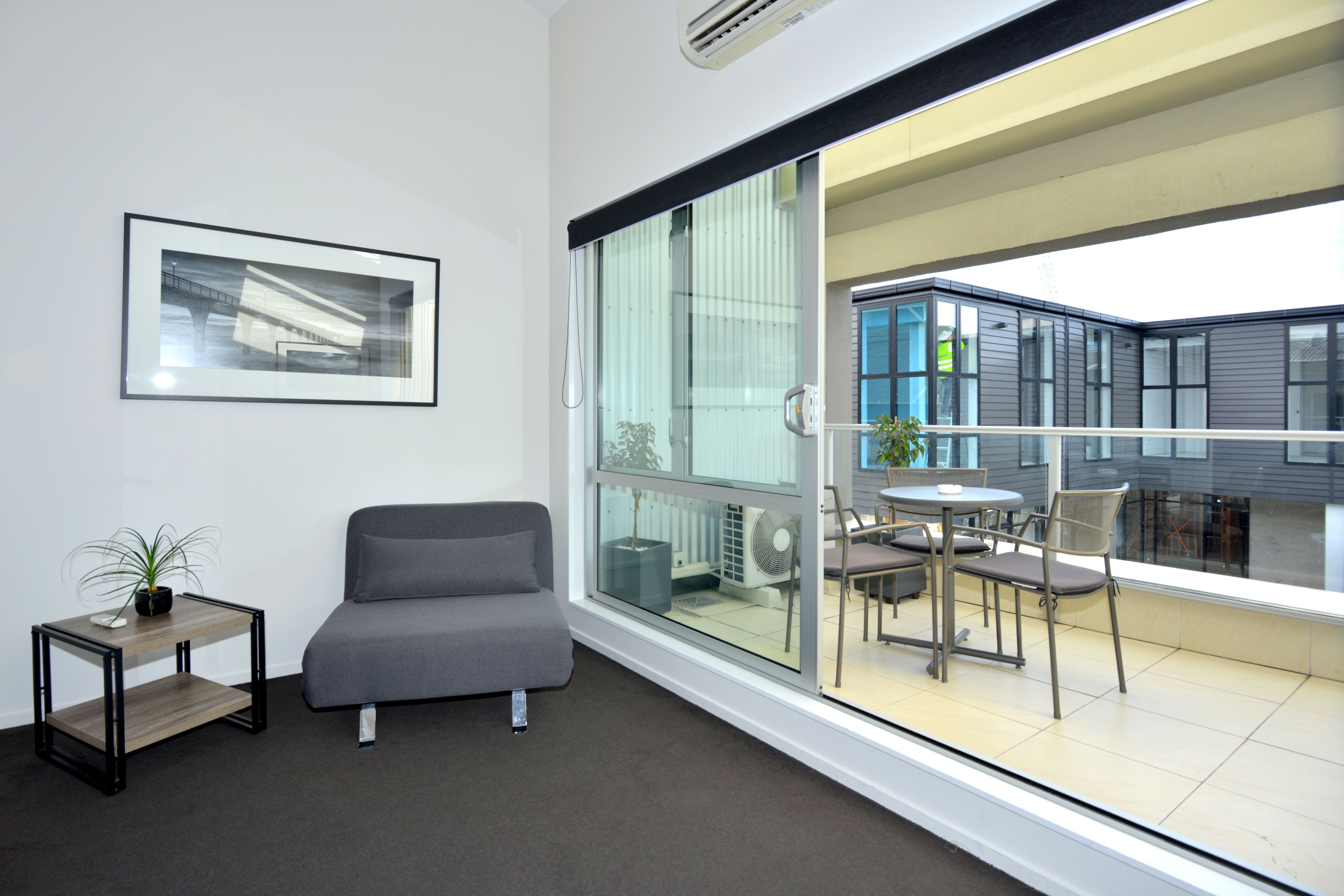 A modern room with a grey sofa, a small table with a plant, and a large sliding glass door leading to an outdoor patio with a table and chairs.