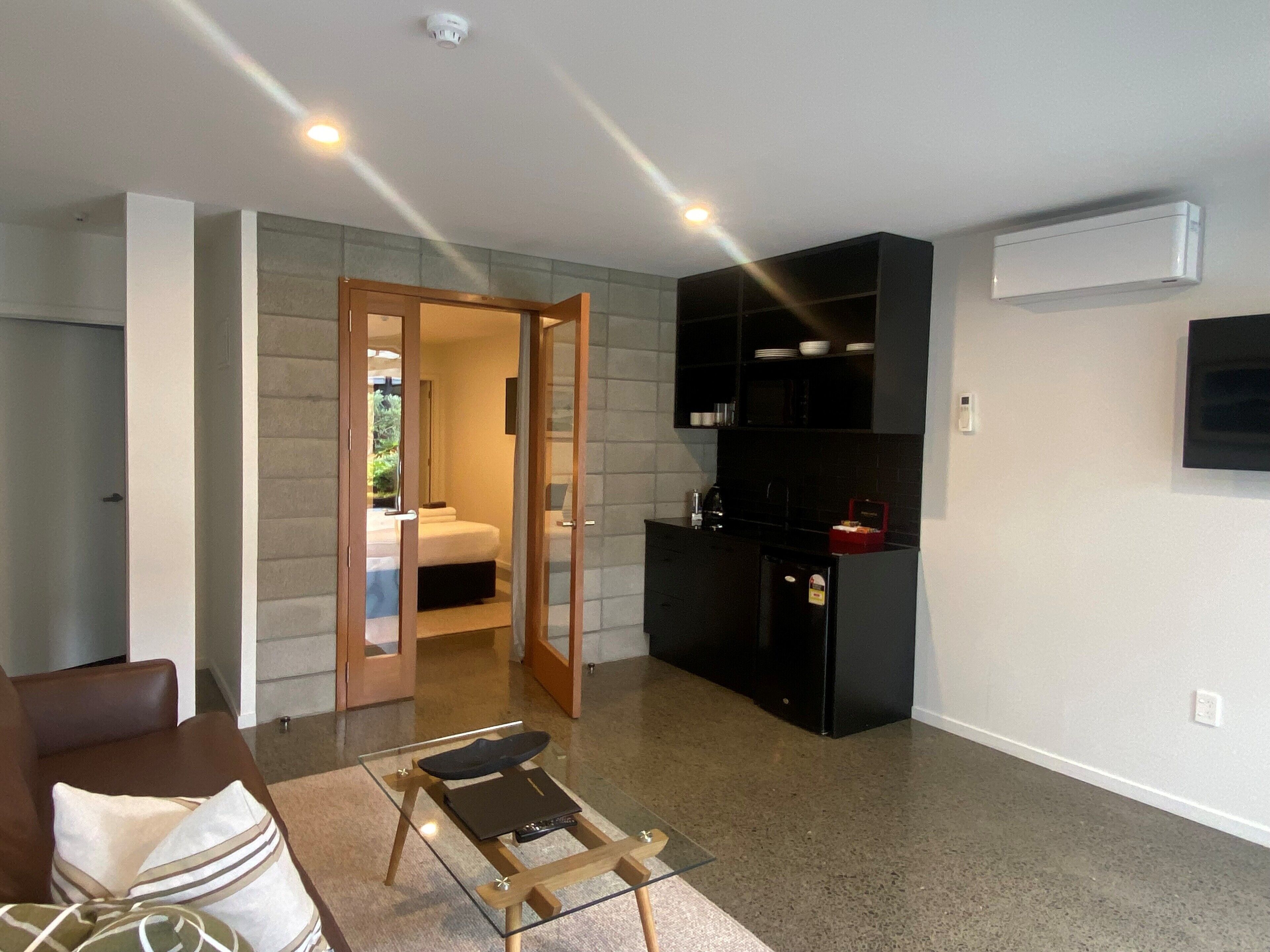 A modern living room with a glass coffee table, a sofa, and a kitchen area with black cabinets and a built-in oven.