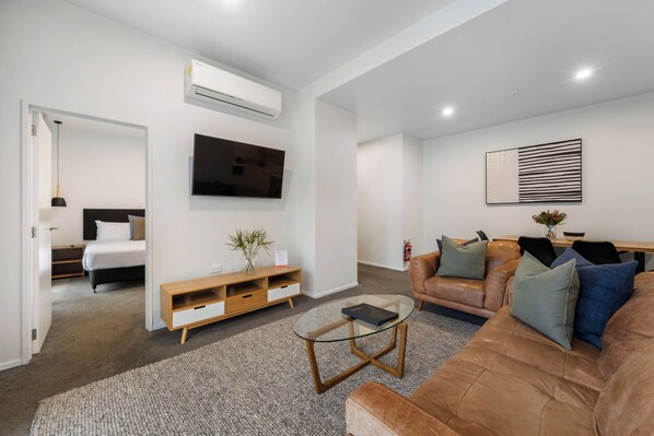 A modern living room with a sofa, a glass coffee table, a TV mounted on the wall, and a small wooden cabinet.